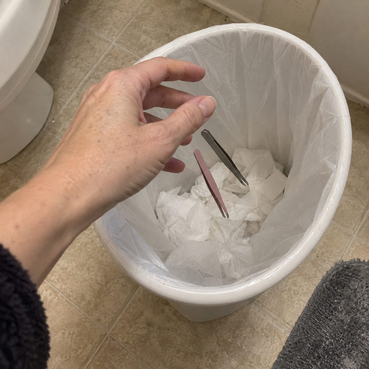 A hand dropping two tweezers into a bathroom bin