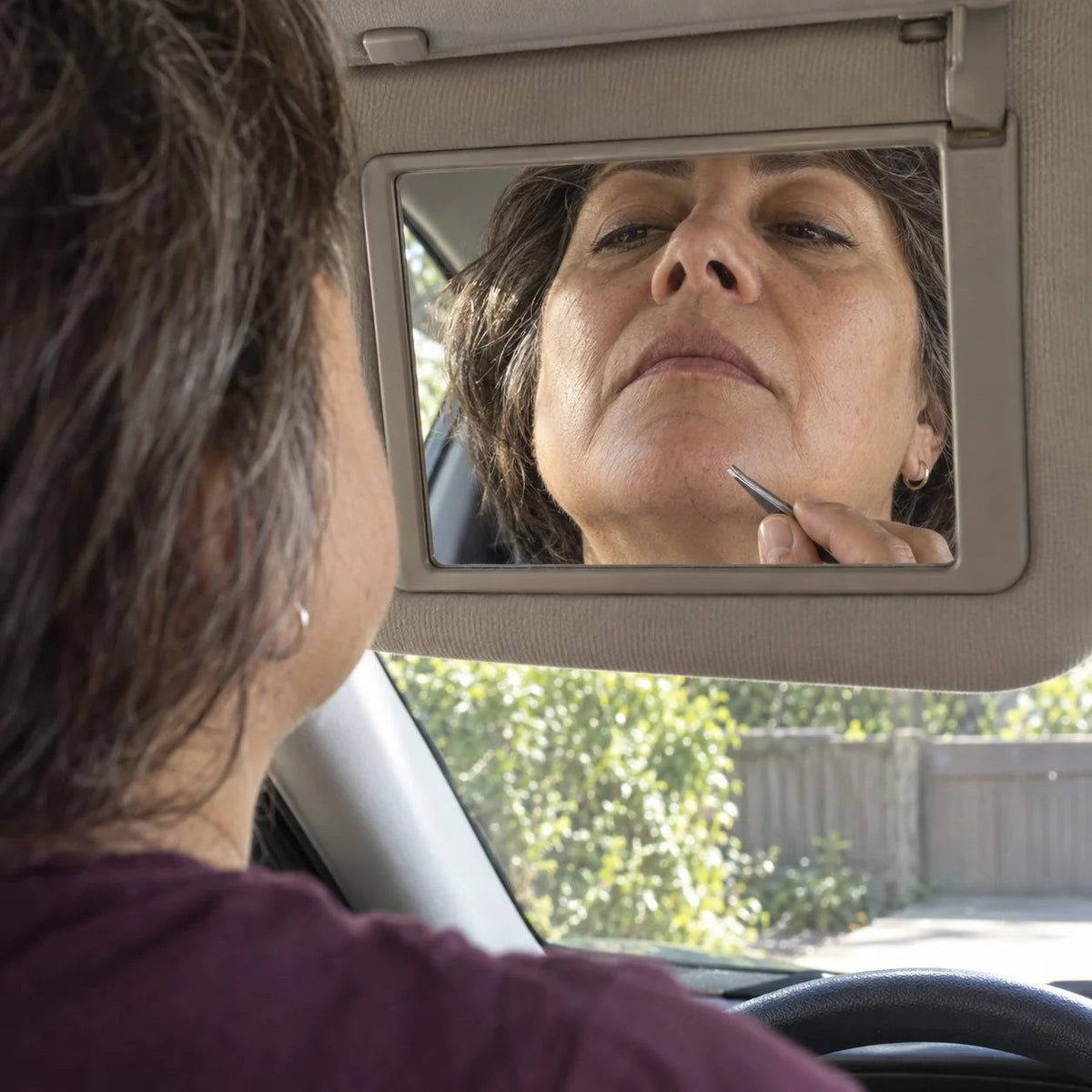 Illustrative parked-car mirror scene of a woman inspecting chin hair with tweezers